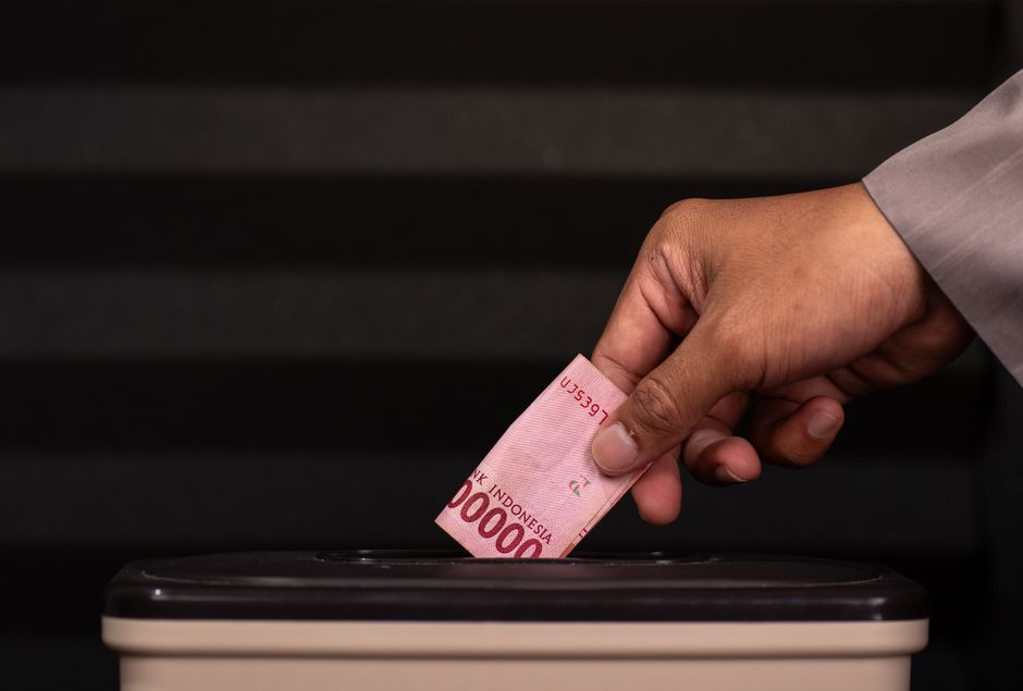 10万インドネシアルピア紙幣を募金箱に入れる手のクローズアップ。慈善活動と寄付を強調している。