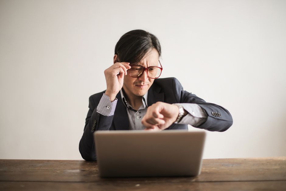 アジア系ビジネスマンがノートパソコンで作業しながら時計を確認し、ストレスと緊急性を表現しています。